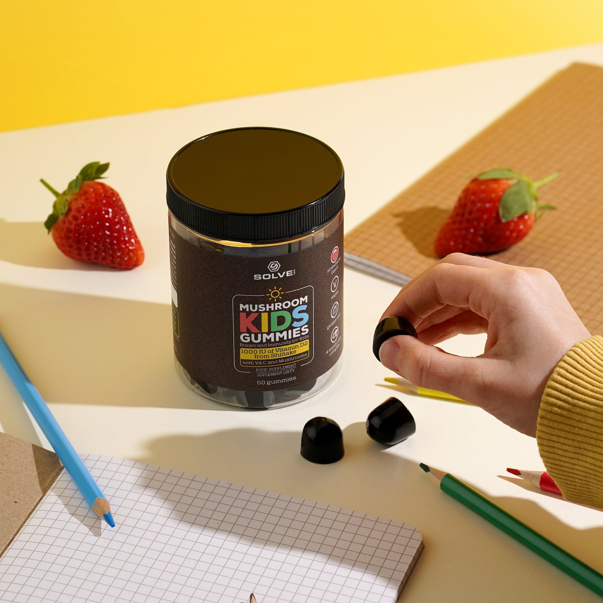 Container of 'Mushroom Kids Gummies' with a hand holding a gummy, surrounded by strawberries and blackberries on a yellow background.